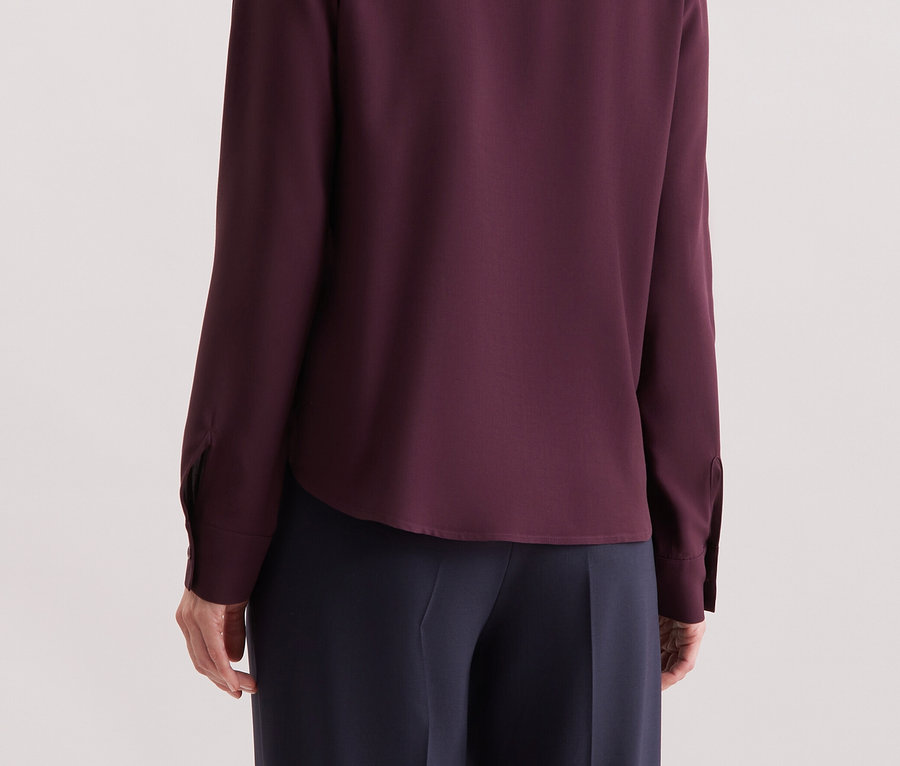 Von hinten ist eine Frau mit blonden Haaren zu sehen, die ein purpurrotes Langarmhemd trägt.