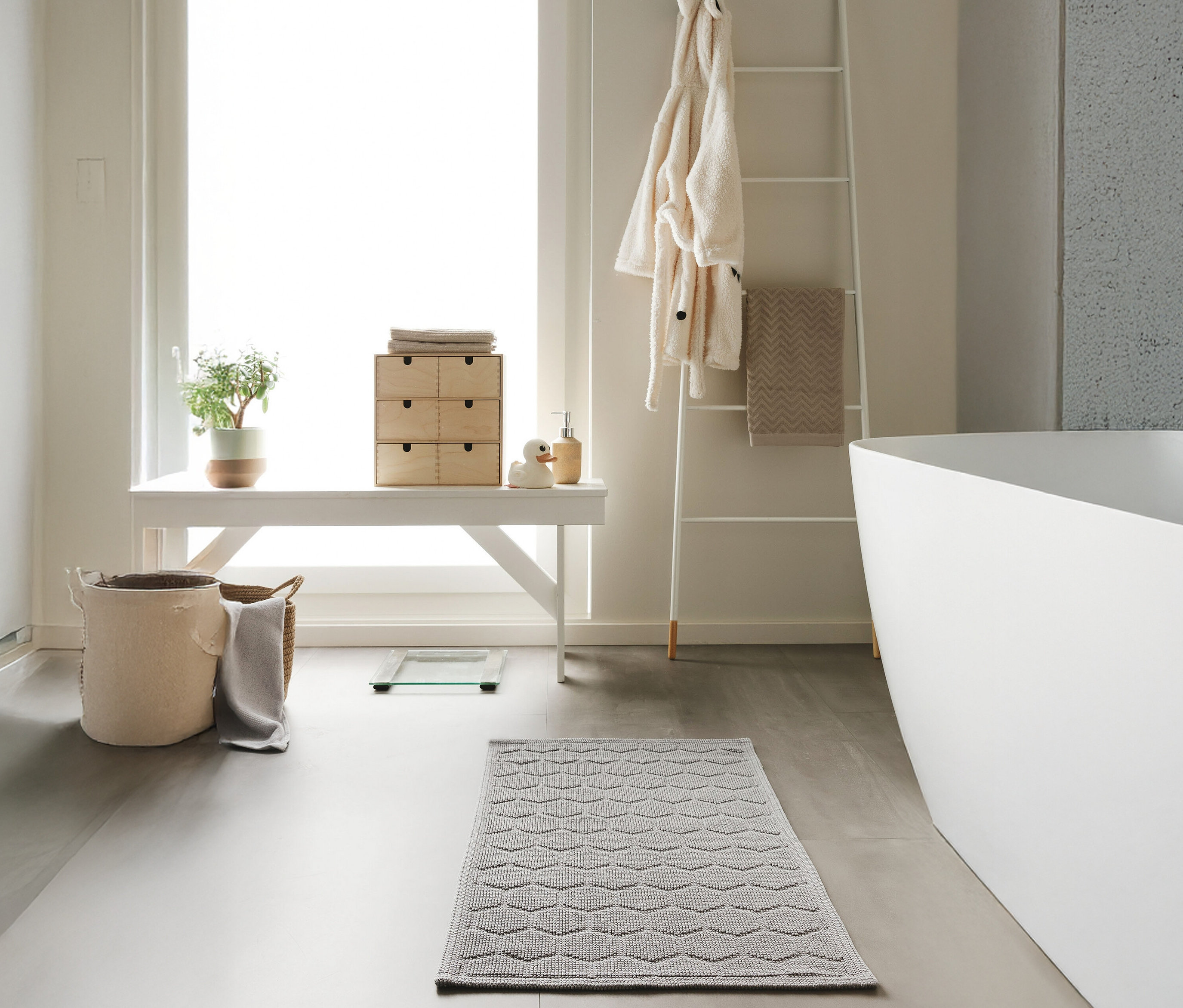 Ein Badezimmer mit einer weißen Bank, einer Leiter und einer Badewanne. Auf dem Boden liegt eine hellgraue Badematte »Jojo« von benuta mit Rautenmuster (50 x 80 cm).