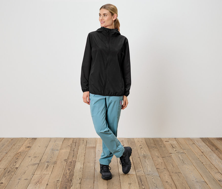 Frau in schwarzer Regenjacke mit Kapuze, hellblauen Hosen und schwarzen Schuhen steht vor einer weißen Wand.