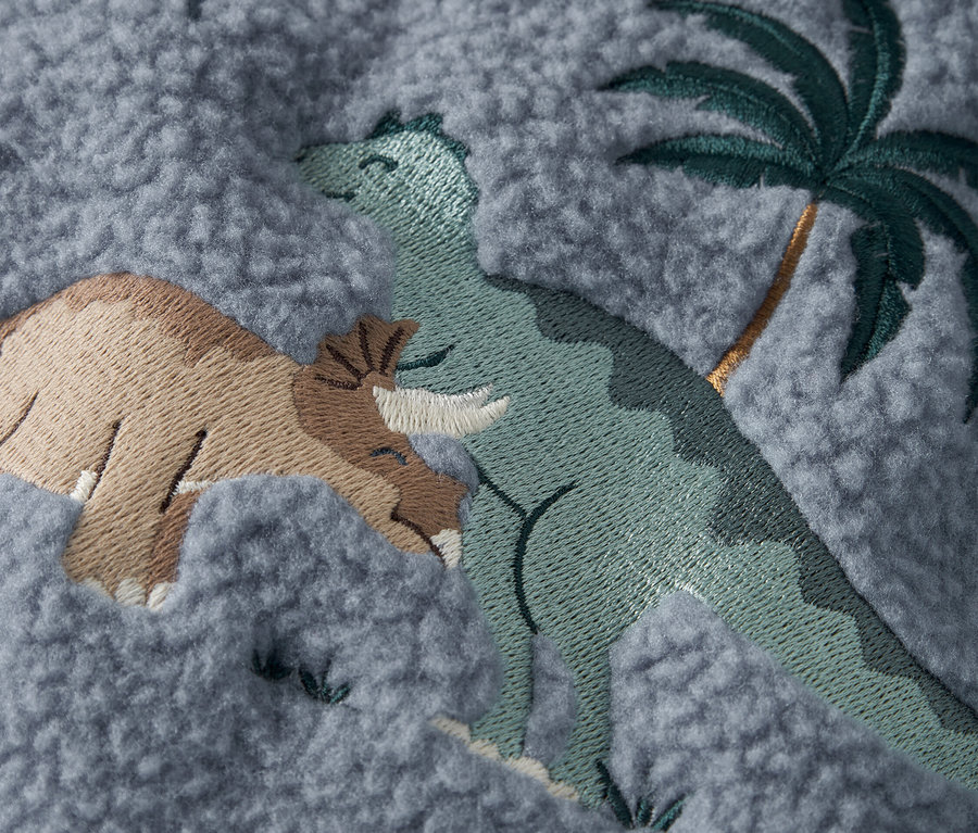 Nahaufnahme eines grauen Stoffes mit aufgestickten Dinosauriern und einer Palme.