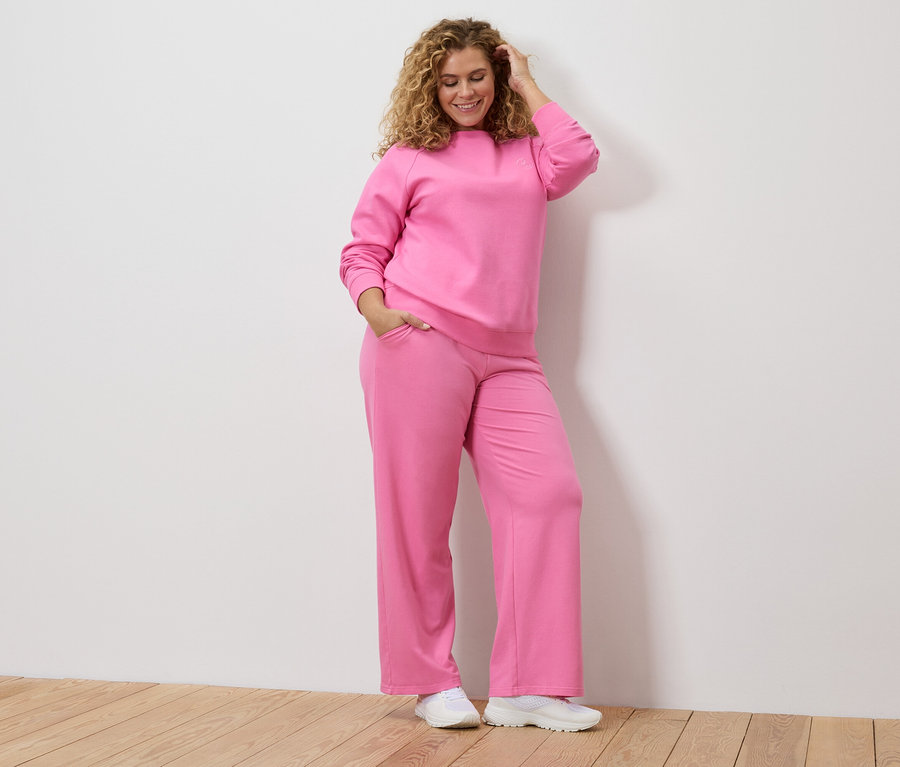 Frau mit lockigem Haar, die ein pinkes Trainingsanzug und weiße Sneaker trägt und ihre Hand auf ihrem Kopf hat.