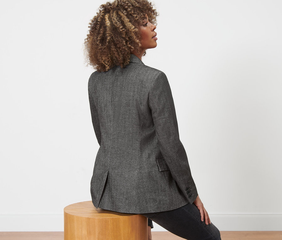 Frau sitzt auf einem Holzhocker und trägt einen Webblazer in Herringbone-Optik.