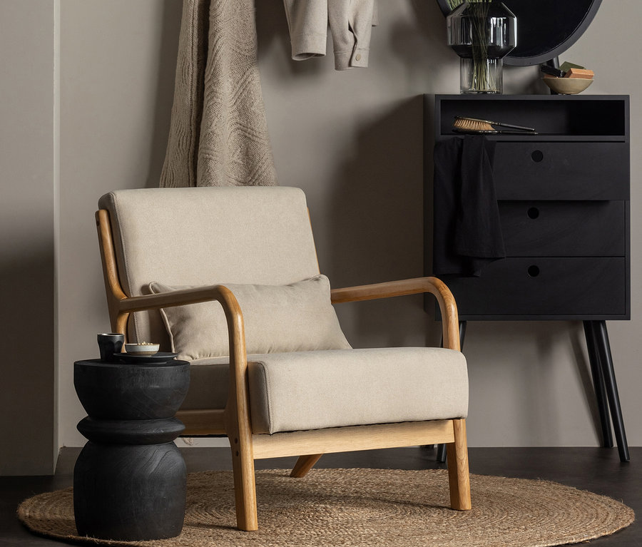 Beiger Sessel mit Kissen steht auf dem naturbelassenen Woood Teppich »Ross« 150 cm. Daneben ein schwarzer Hocker, dahinter eine schwarze Kommode und ein runder Spiegel.