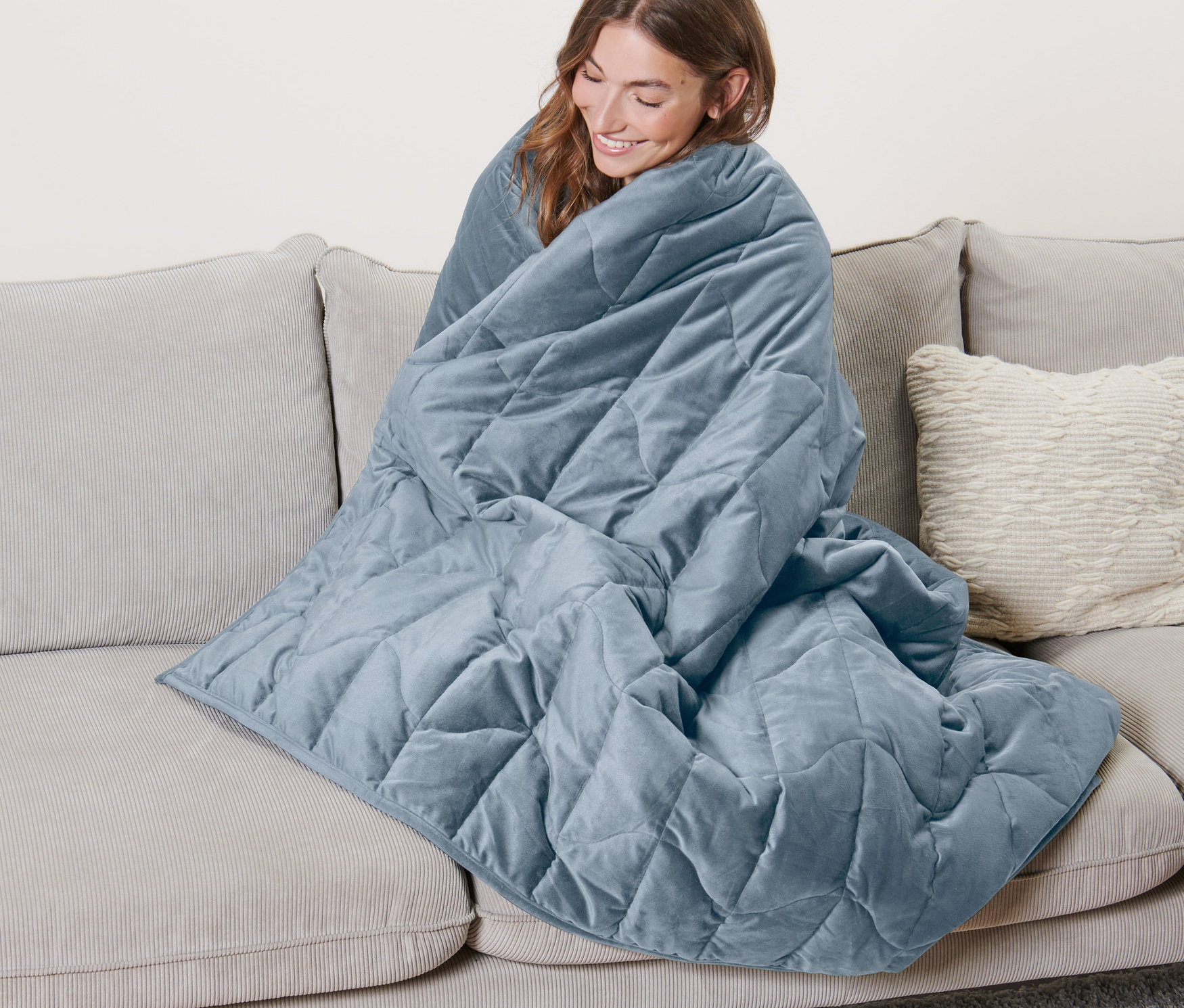 Frau sitzt auf Sofa, eingewickelt in eine blaue Entspannungs-Gewichtsdecke, 8 kg.