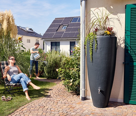 Eine Frau sitzt im Stuhl, ein Mann bewässert den Garten und rechts steht ein graphitgraues Regenfass.