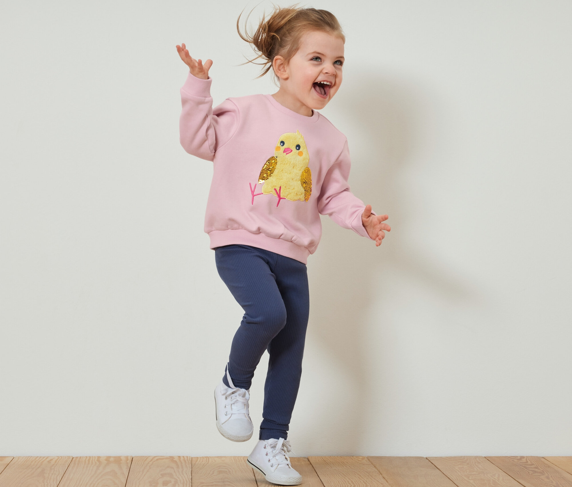 Mädchen springt in einem Kinder-Sweatshirt mit angerauter Innenseite und Leggings auf einem Holzboden.