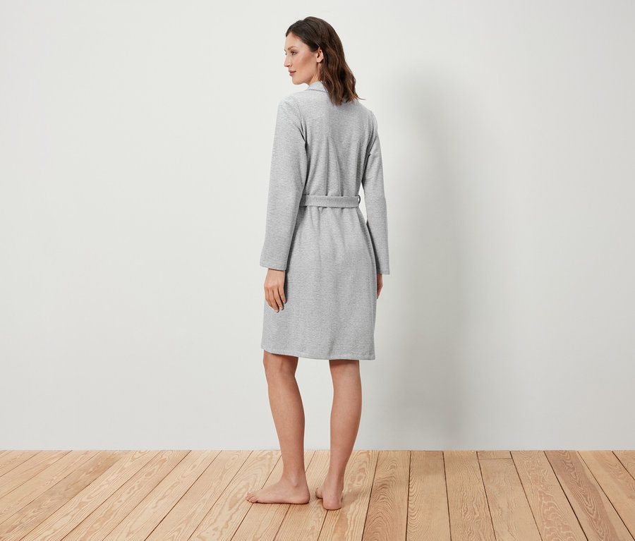 Frau steht mit dem Rücken zum Betrachter und trägt einen grauen Bademantel.