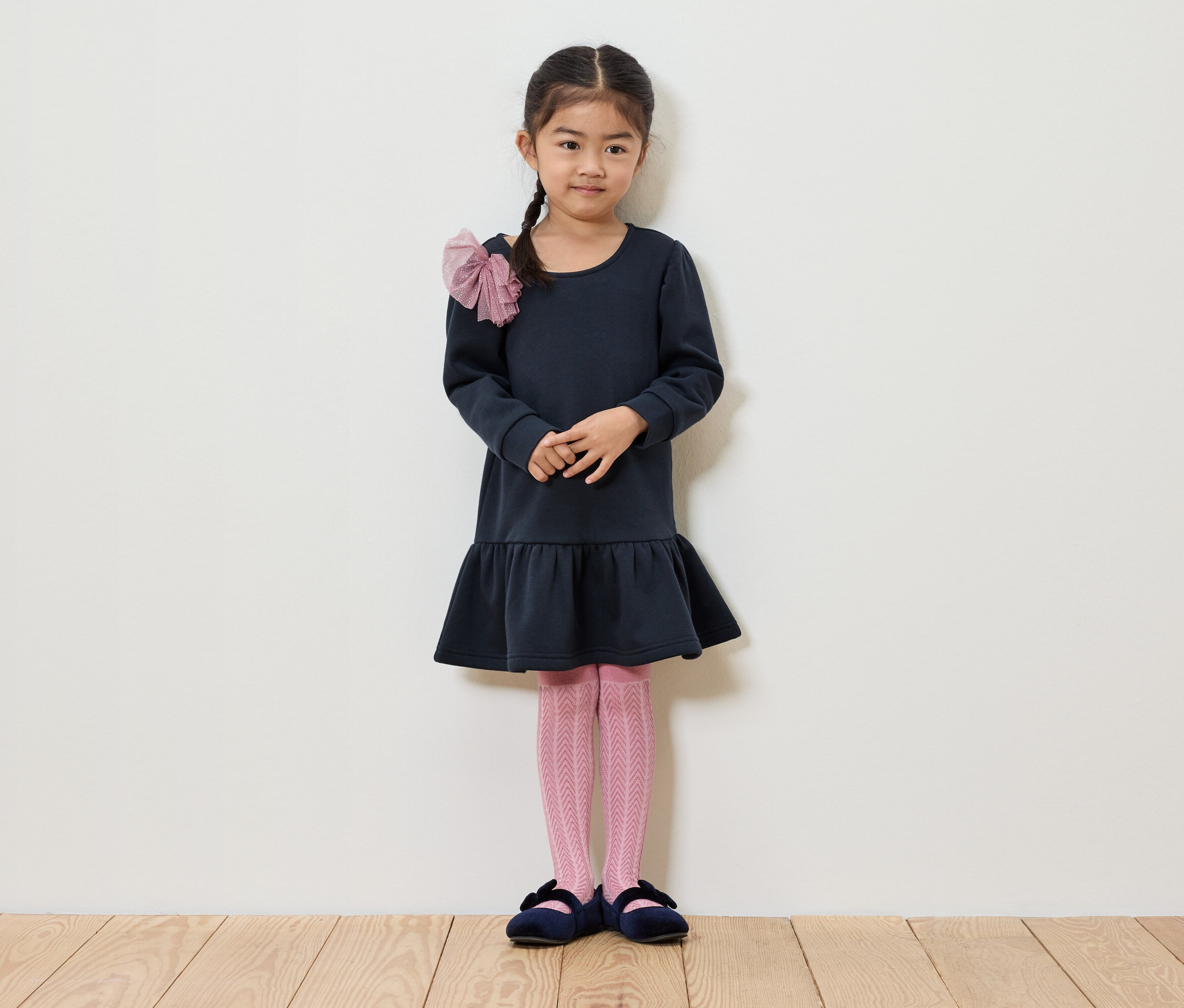 Ein Mädchen mit Zopf steht vor einer weißen Wand. Sie trägt ein dunkelblaues Kleid, eine rosa Strumpfhose und blaue Schuhe.