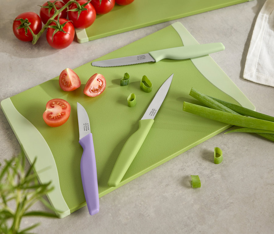 Grünes Schneidebrett mit geschnittenen Tomaten, Frühlingszwiebeln und drei Messern mit farbigen Griffen.