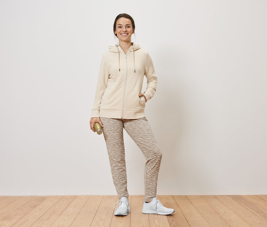 Frau steht und hält eine goldene Flasche. Sie trägt eine beige Reißverschlussjacke mit Kapuze, gemusterte Hosen und weiße Turnschuhe.