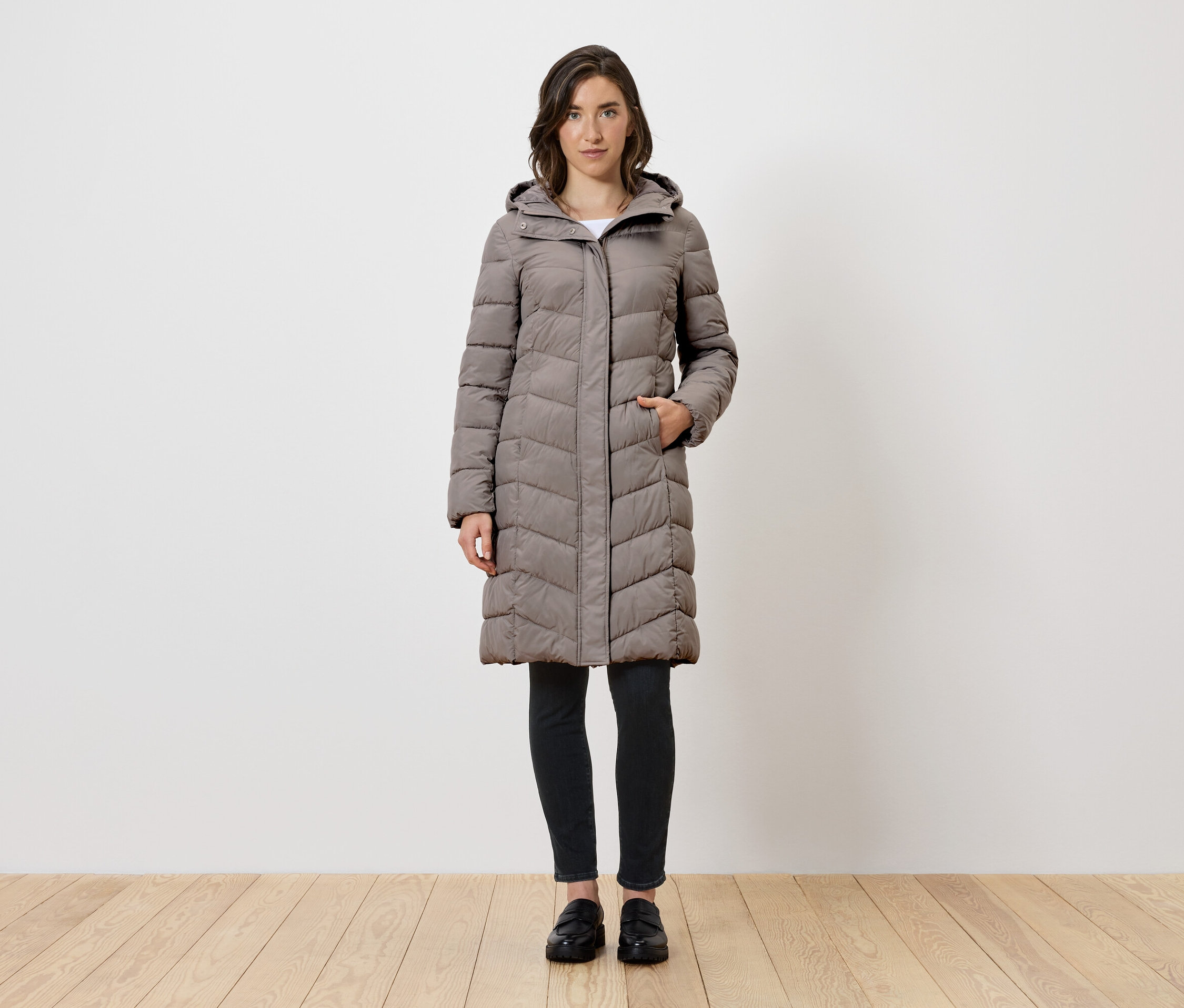 Frau steht und trägt einen langen, grauen Daunenmantel mit Kapuze, schwarze Leggings und schwarze Schuhe.