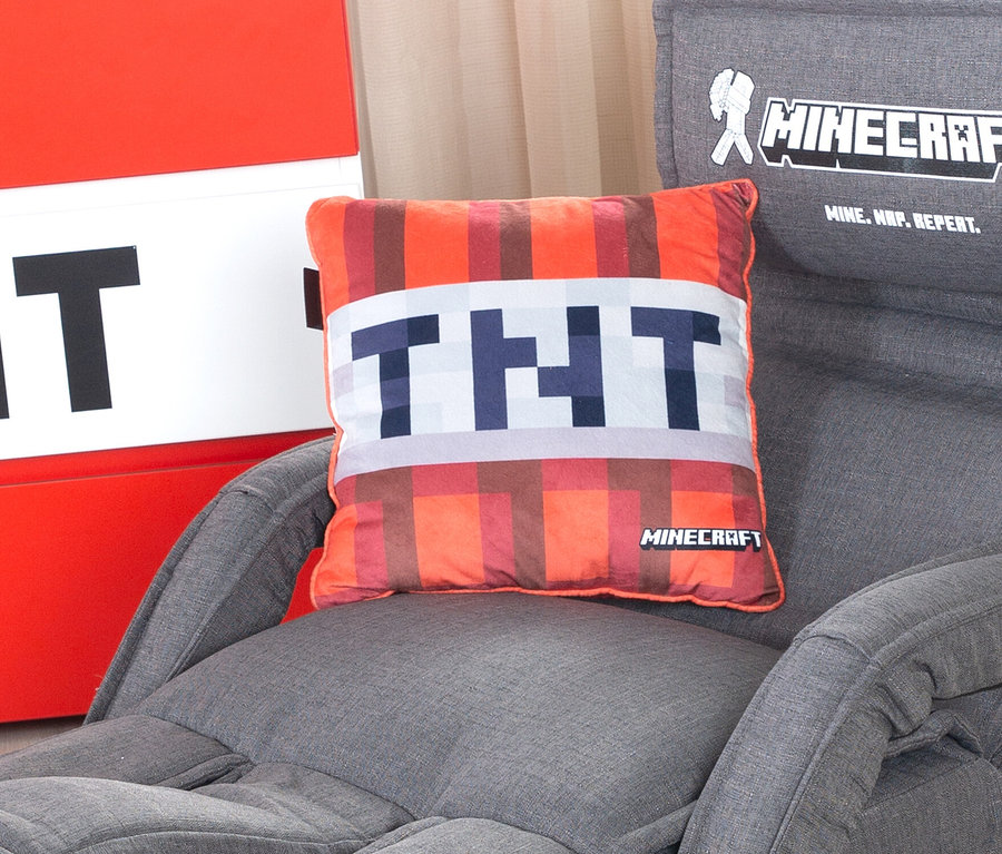 Ein Minecraft TNT Dekokissen liegt auf einem grauen Sessel. Im Hintergrund ist eine rot-weisse Aufbewahrungsbox zu sehen.