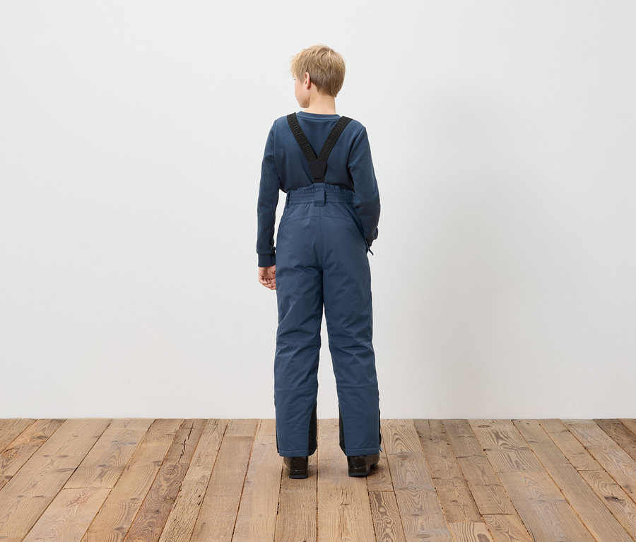 Junge steht mit dem Rücken zur Kamera und trägt blaue Latzhose.