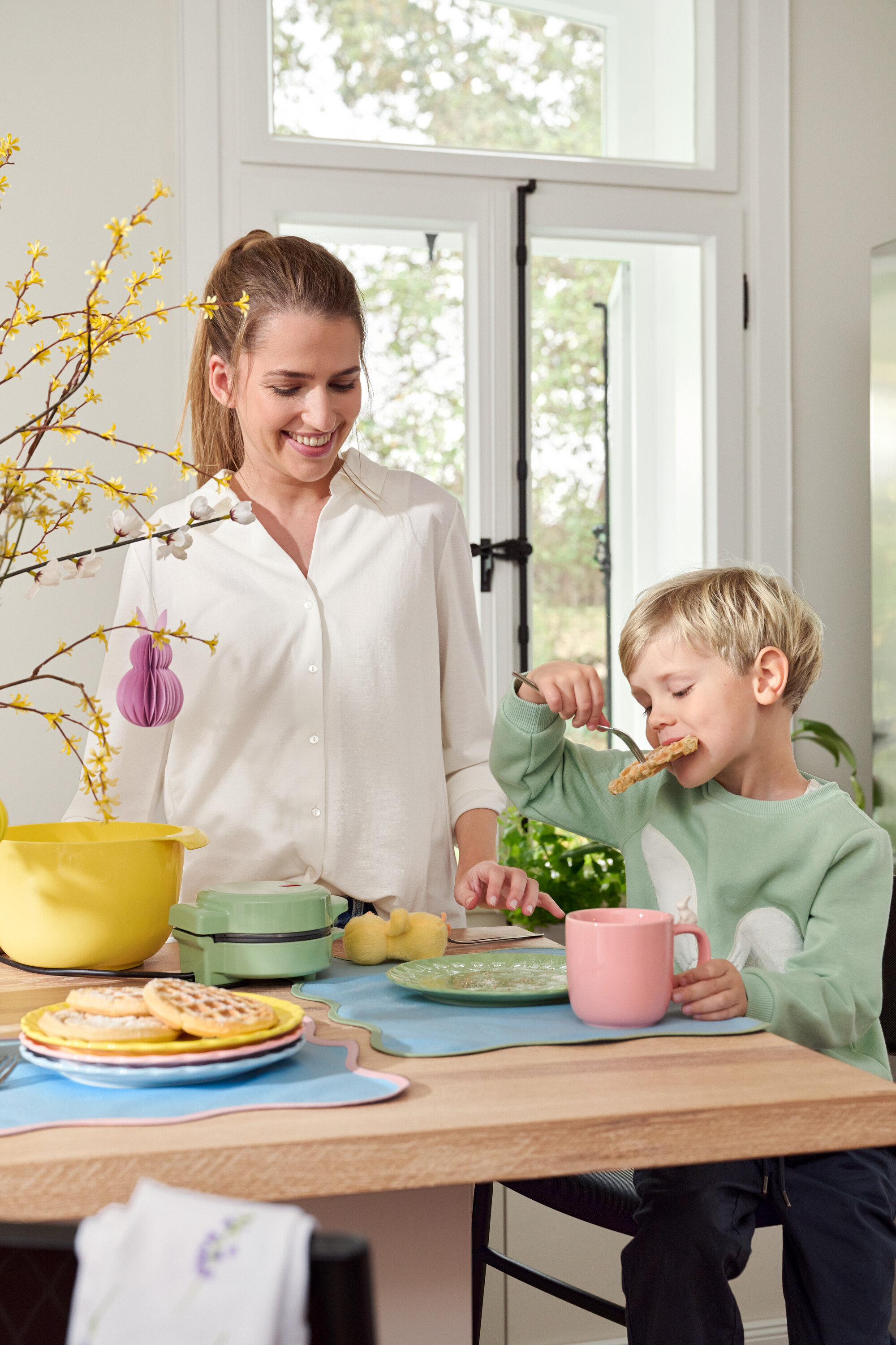 Frau und Kind am Tisch. Kind isst Waffel mit Gabel, Frau lächelt.