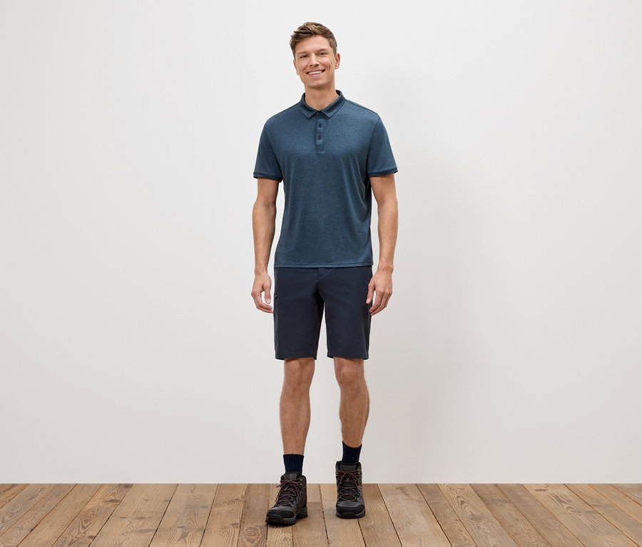 Ein Mann steht vor einer weißen Wand und trägt ein blaues Poloshirt, kurze Shorts und Wanderschuhe.