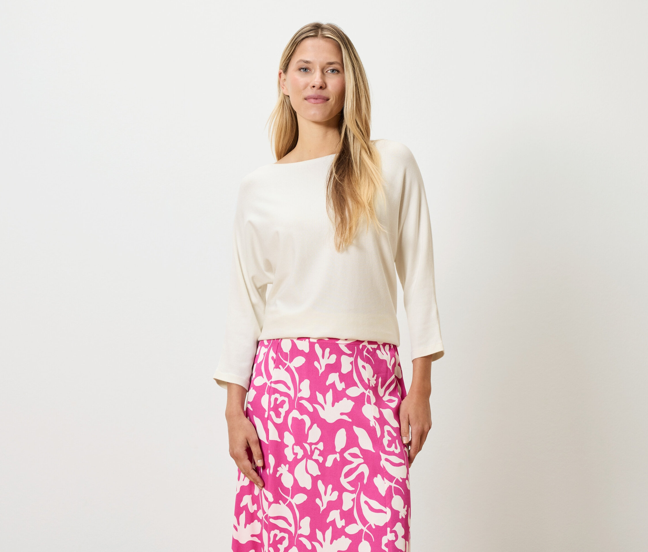 Eine Frau mit blonden Haaren trägt ein cremefarbenes Oberteil und einen pinkfarbenen Rock mit Blumendruck.