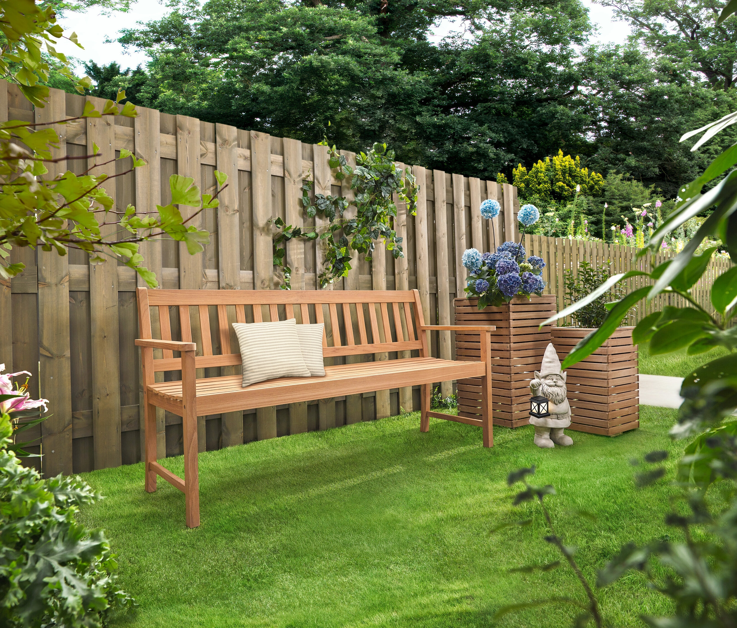 Eine Holzbank mit Kissen steht auf einem Rasen. Dahinter stehen zwei Pflanzsäulen »Tinus« mit blauen Blumen.