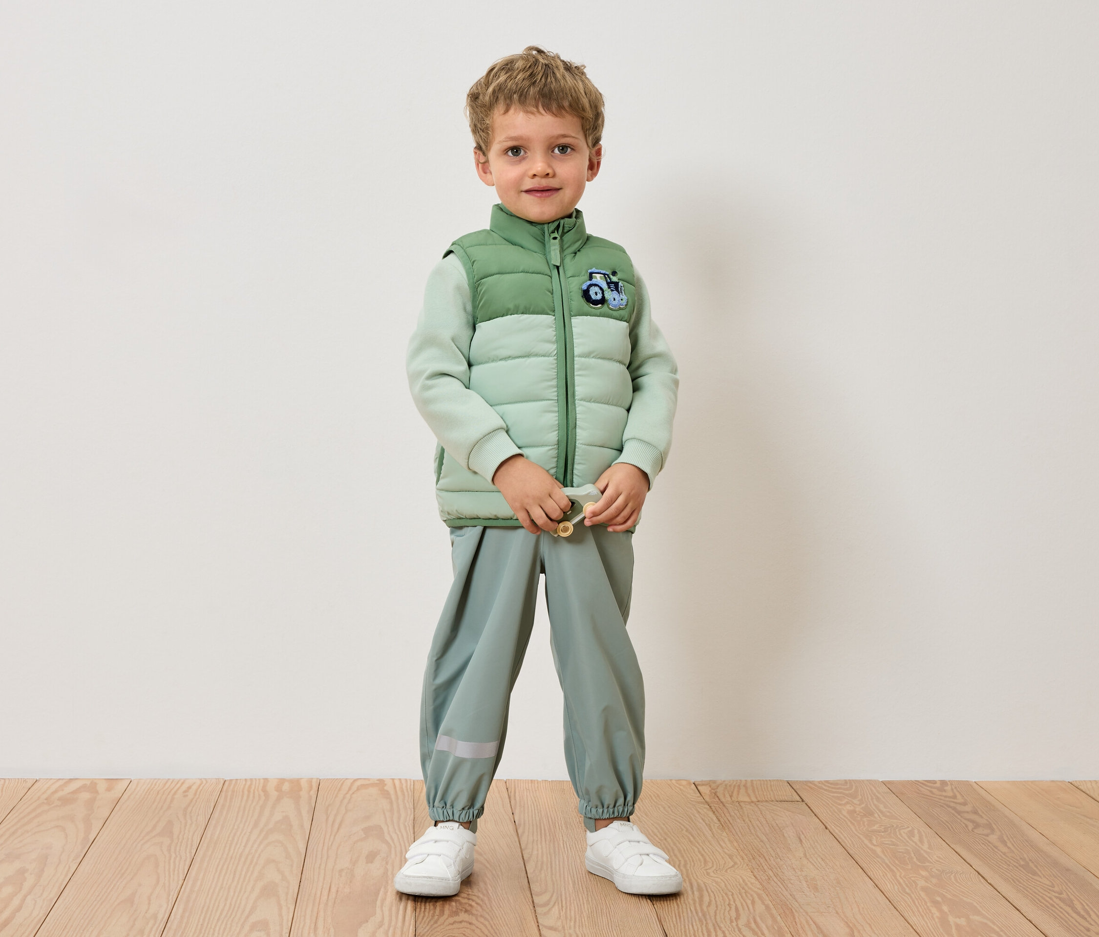 Ein Junge steht in einer hellgrünen Jacke, einer hellgrünen Hose und weißen Turnschuhen vor einer Wand.