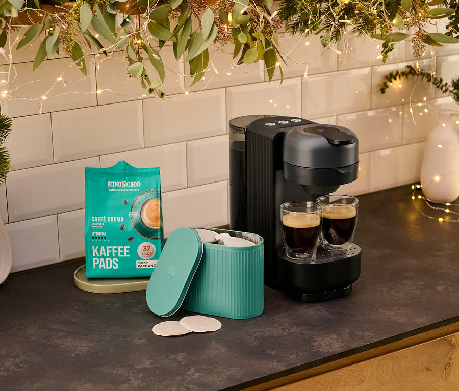 Eine »Call me Pad« Kaffee Padmaschine Graphite steht neben zwei Gläsern Kaffee. Daneben befindet sich eine Packung Kaffeepads und eine Dose mit Kaffeepads.