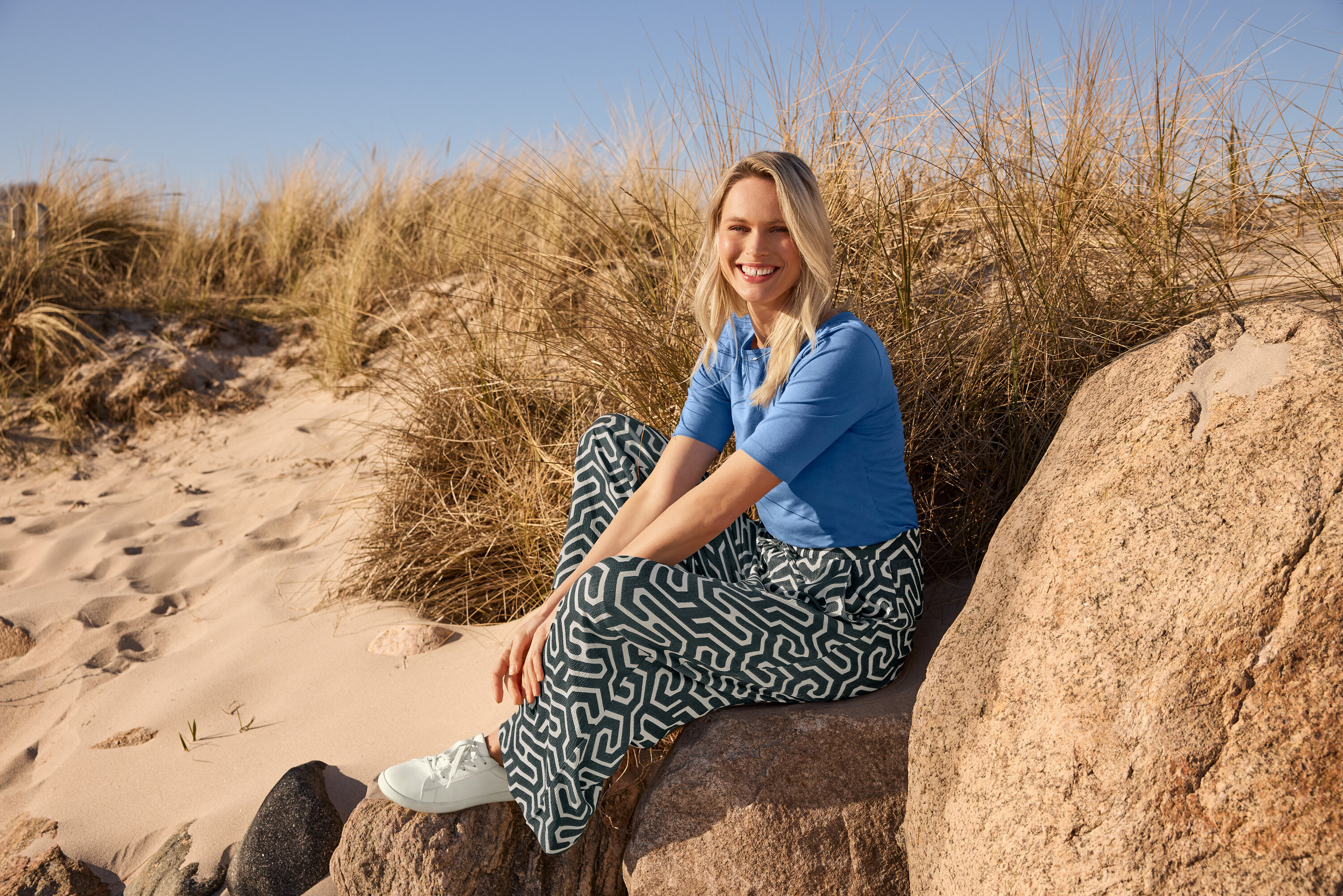 Eine Frau sitzt am Strand auf einem Stein und trägt ein blaues Halbarmshirt, eine bedruckte Schlupfhose in Crinkle-Optik und Ledersneaker.