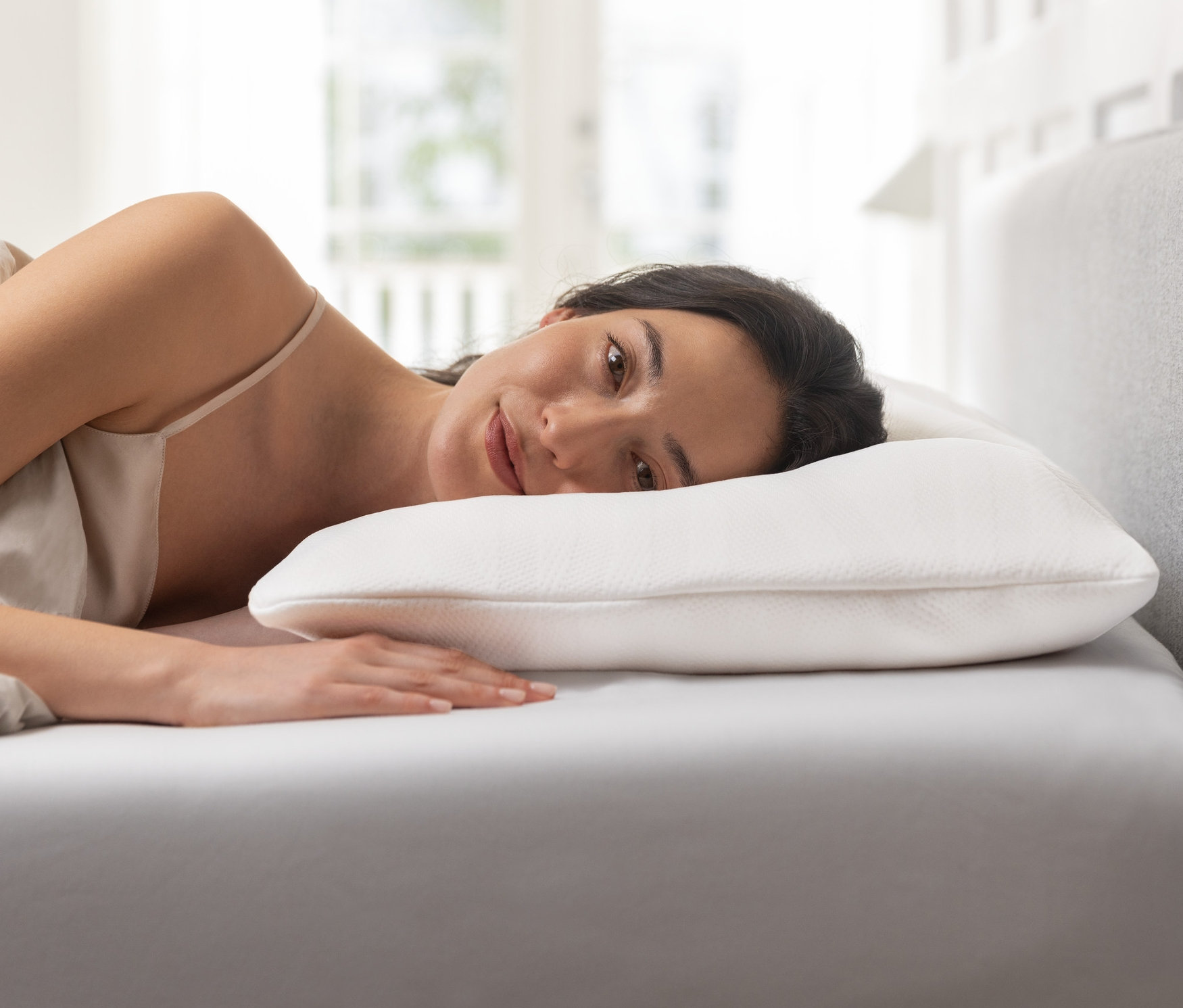 Frau liegt seitlich auf einem weißen Bettlaken und lächelt, ihr Kopf ruht auf einem TEMPUR® Comfort Pureclean Schlafkissen.