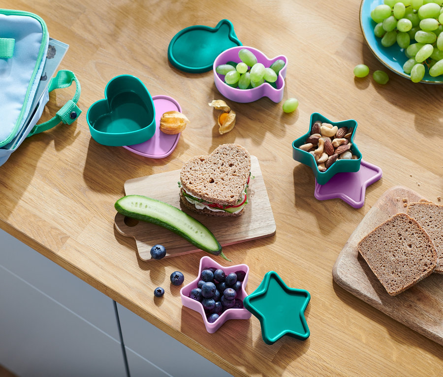 Auf einem Holztisch liegt ein Snack. Herzförmiges Sandwich, Gurke, Blaubeeren in einem sternförmigen Behälter, Nüsse, Trauben in einem fischförmigen Behälter und Brot.