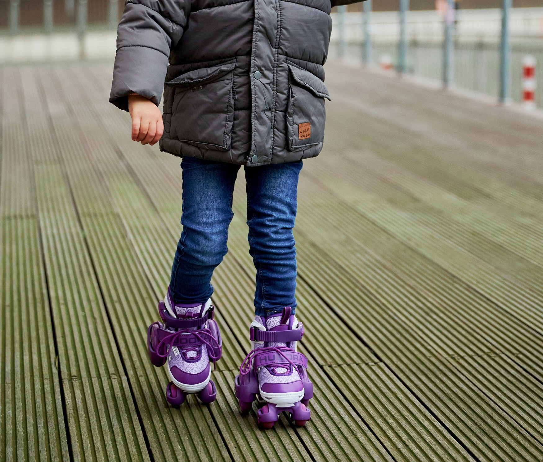 Mädchen in grauer Jacke und lila HUDORA Rollschuhe »My First Quad« steht auf Holzdeck und hält Händchen mit einem Erwachsenen.
