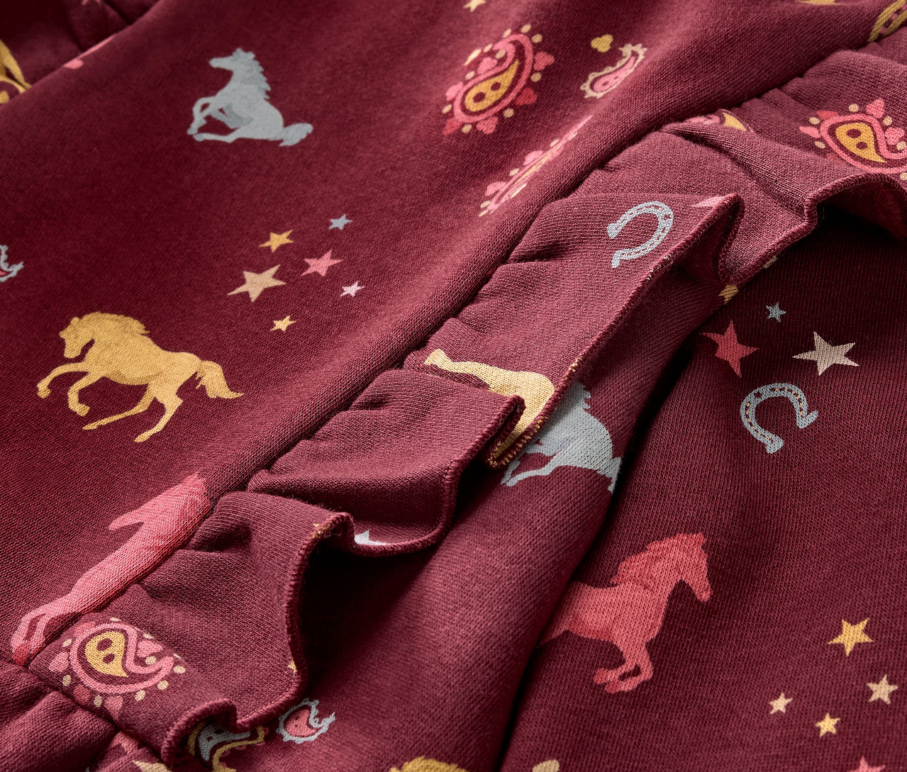 Detailaufnahme eines bordeauxroten Kinder-Sweatkleides mit Rüschen und Pferde-, Stern- und Hufeisenmuster.
