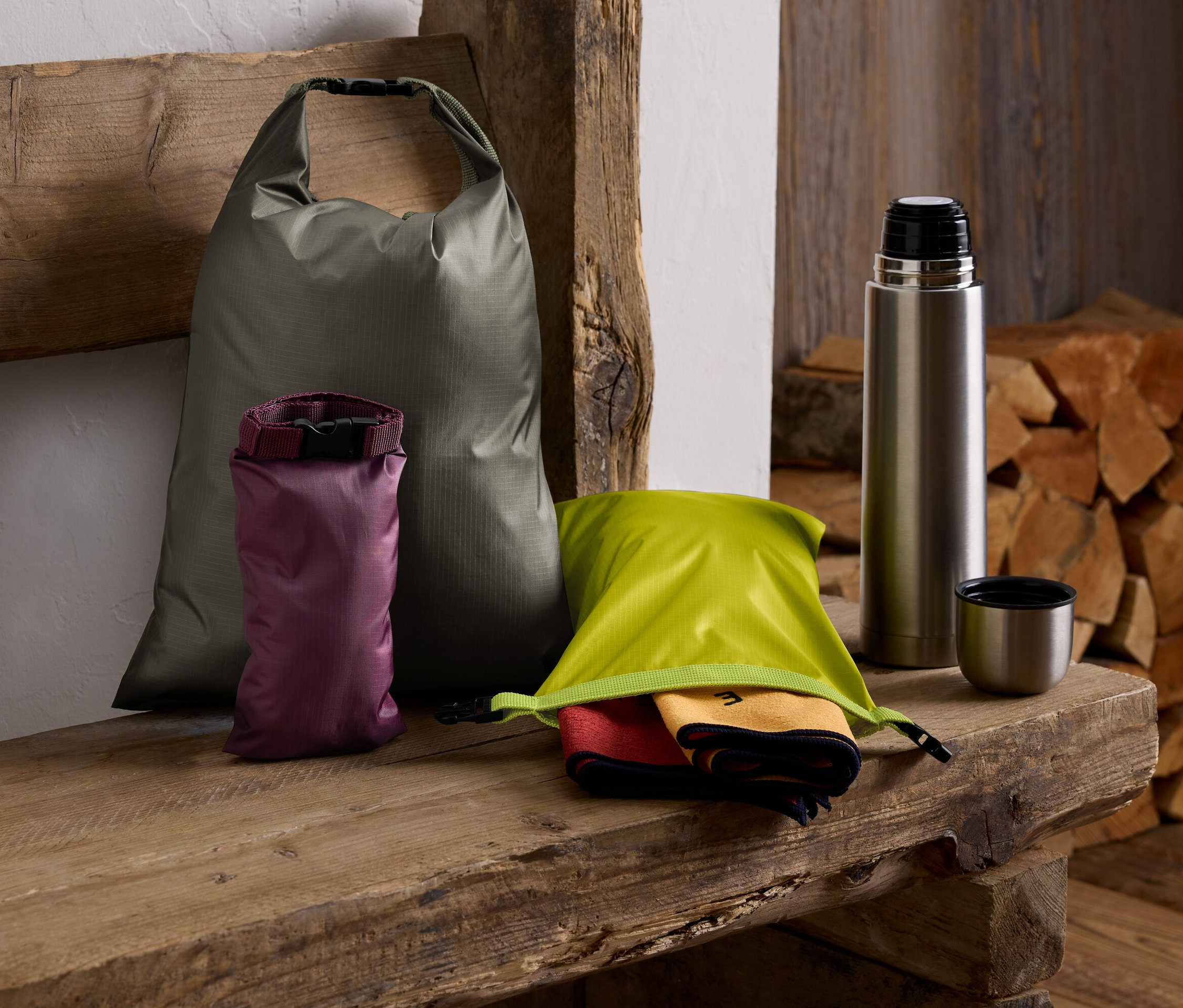 Auf einer Holzbank liegen ein grüner Sack, ein lila Sack, ein grüner Sack mit Tüchern, eine Thermoskanne und ein Holzstapel.