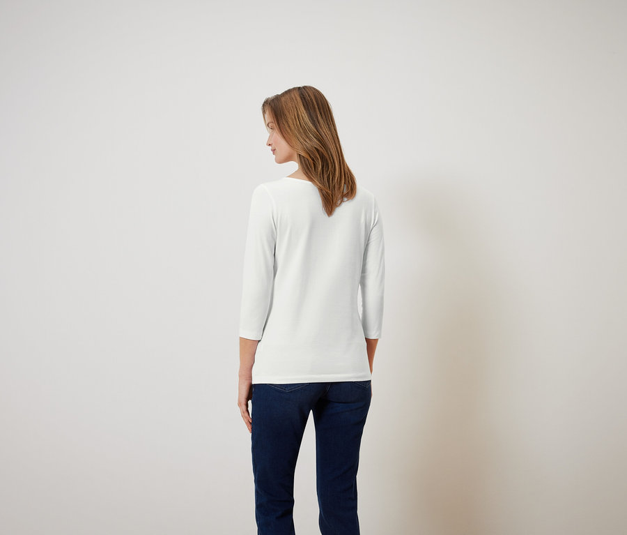 Frau mit braunen Haaren steht mit dem Rücken zur Kamera und trägt ein weißes Dreiviertelarmshirt und blaue Jeans.