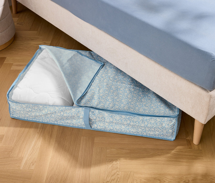 Unter einem Bett liegt eine blaue Aufbewahrungstasche mit weißer Füllung.