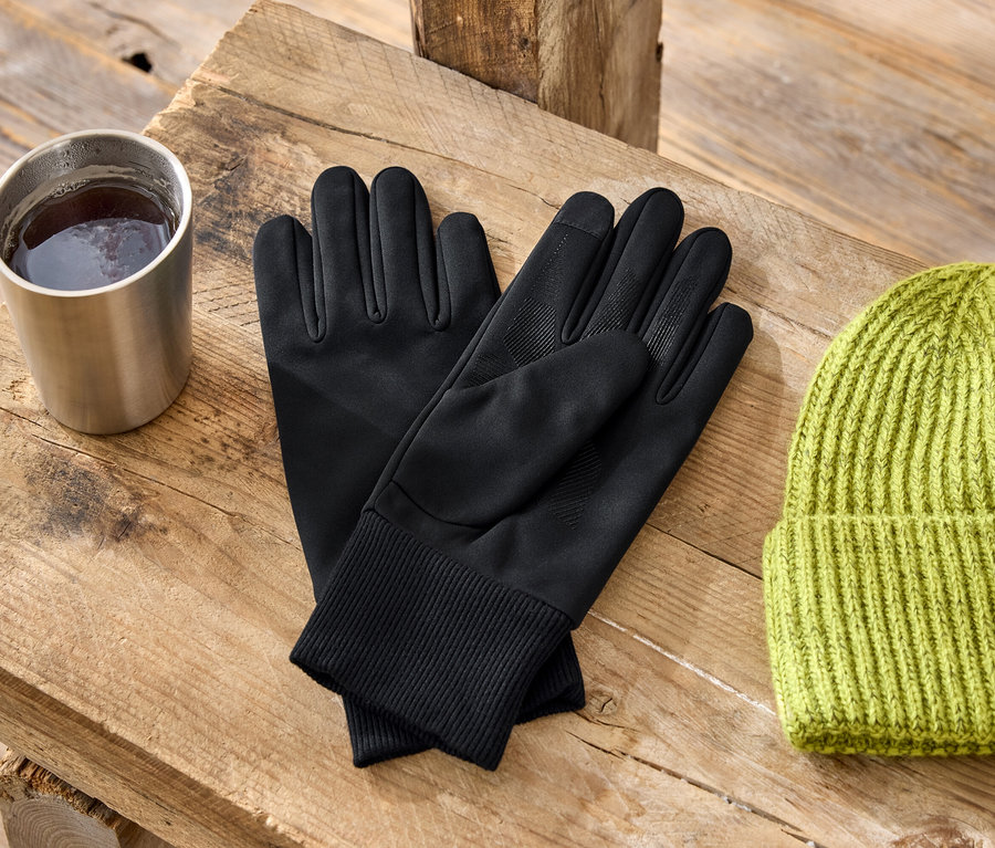 Auf einem Holztisch stehen ein Metallbecher, schwarze Handschuhe und eine grüne Strickmütze.