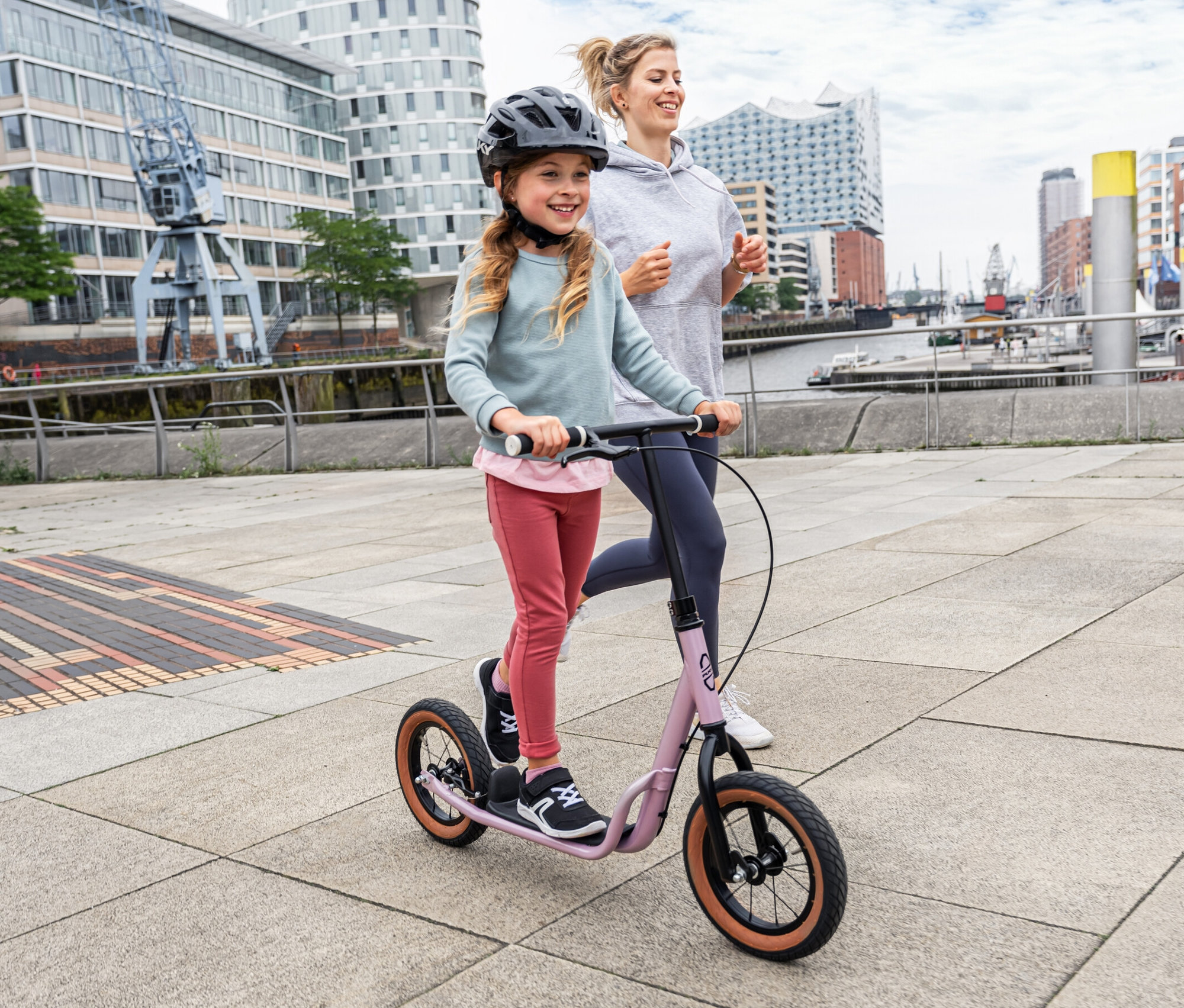 Mädchen fährt Puky Scooter R 07 L, pearl pink, während eine Frau auf dem Gehweg läuft.