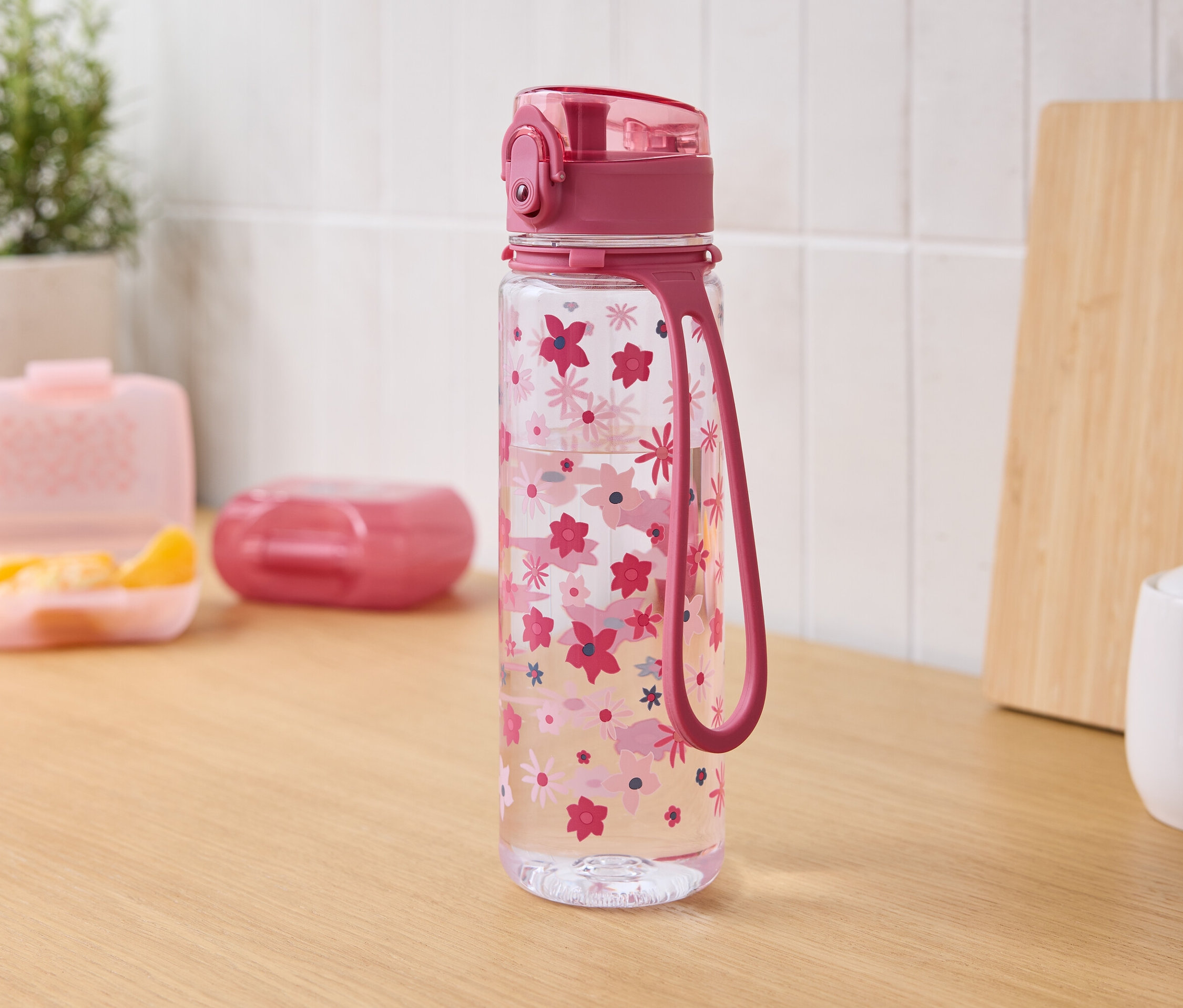 Auf einem Holztisch stehen eine durchsichtige Wasserflasche mit rosa Blüten, rosa Deckel und Schlaufe. Im Hintergrund befinden sich eine Brotdose mit Mandarinen, eine geschlossene Dose, ein Schneidebrett und eine Zuckerdose.