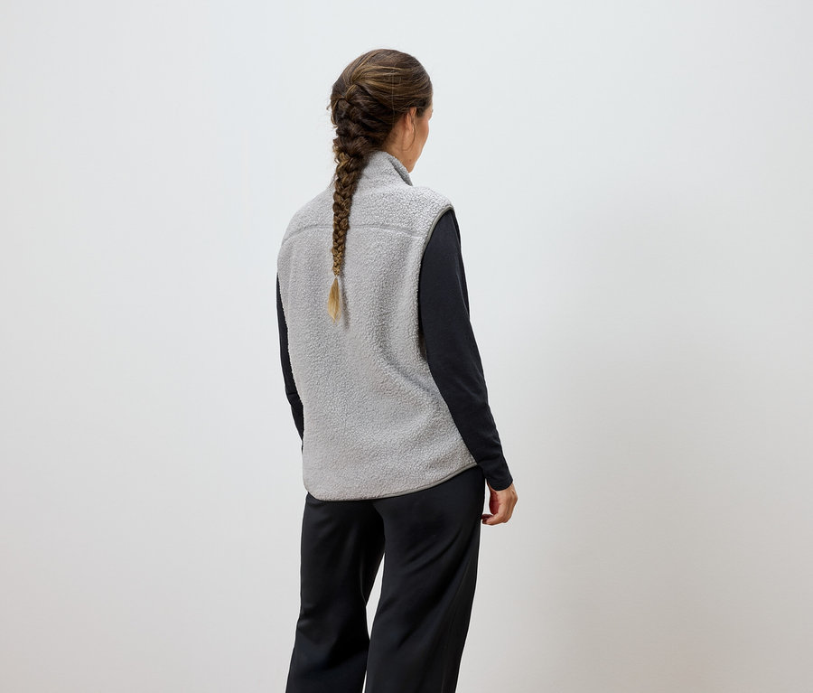Frau mit Zopf von hinten, trägt eine graue Weste und eine schwarze Hose.