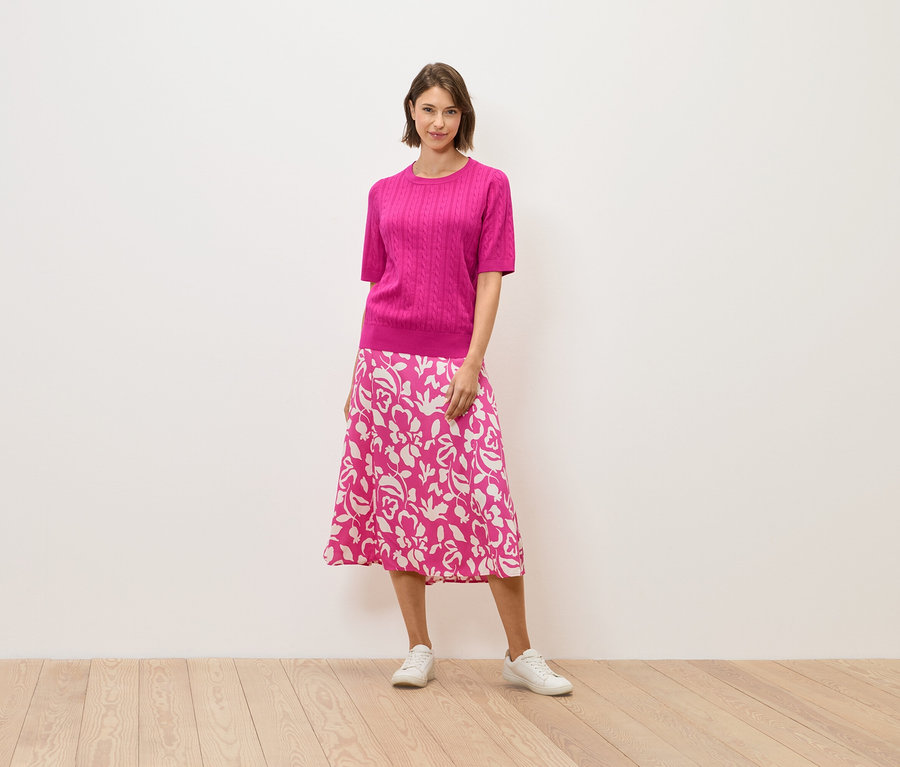 Eine Frau in einem rosa Pullover und einem Rock mit Blumenmuster steht auf einem Holzboden.