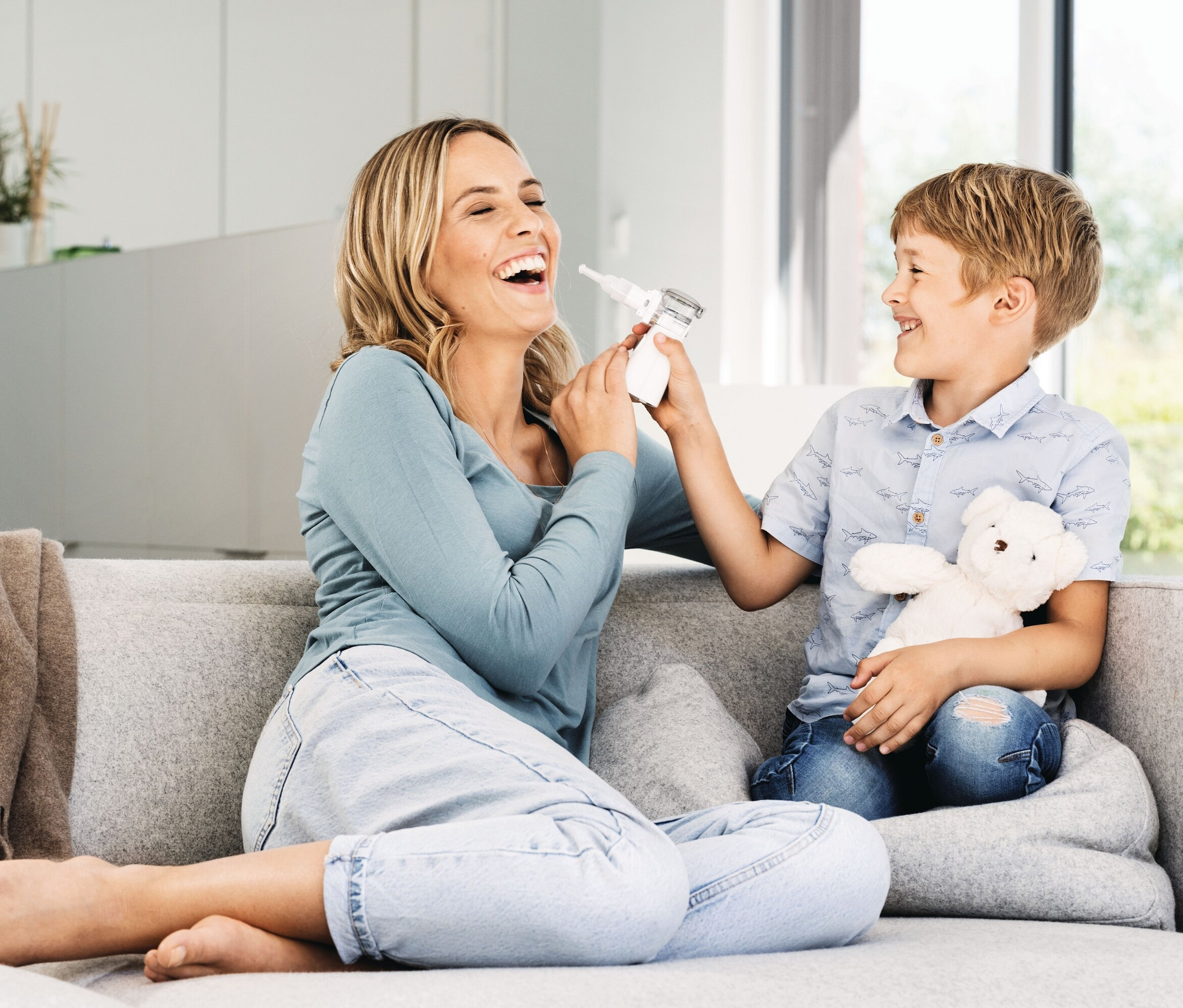 Eine lachende Frau sitzt barfuß auf einem grauen Sofa, während ein Junge ihr ein Nasenreinigungsgerät ins Gesicht hält.