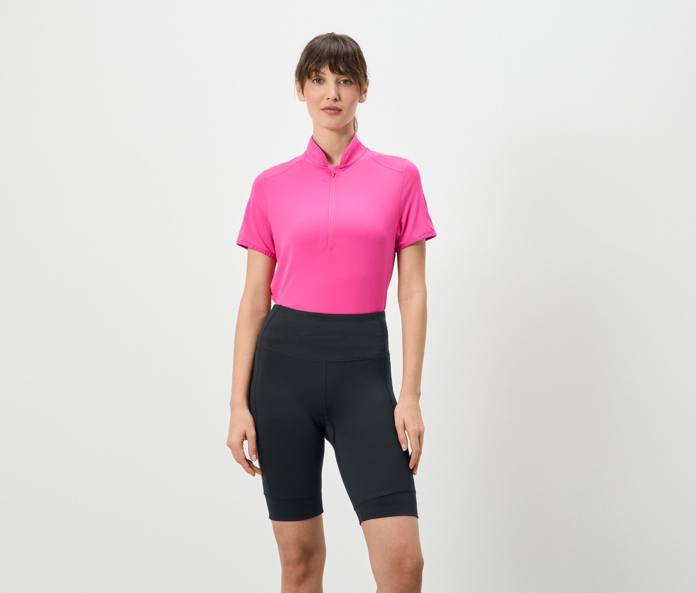 Frau steht und trägt ein pinkes Shirt mit kurzem Reißverschluss und kurze, dunkle Leggings vor einem weißen Hintergrund.