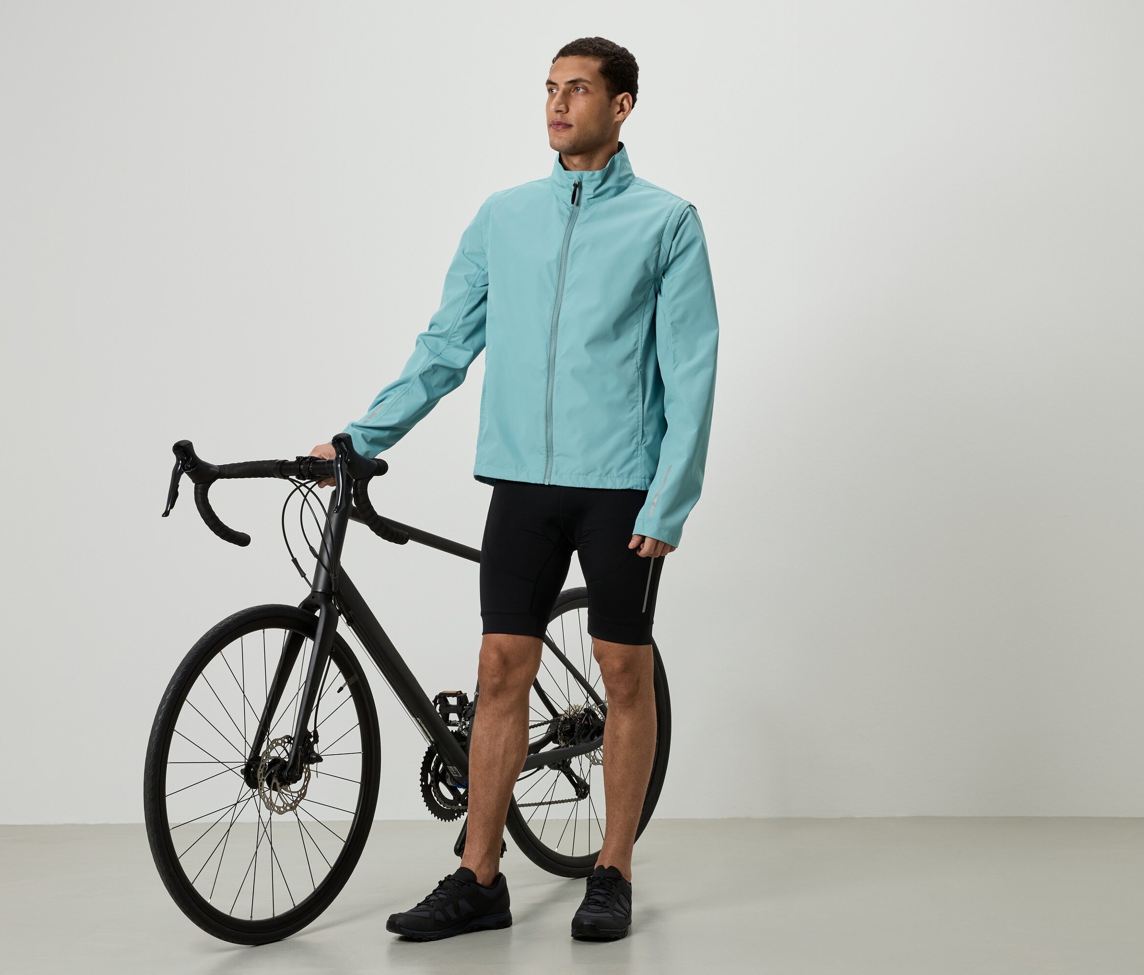 Ein Mann in hellblauer Jacke und schwarzen Shorts steht neben einem schwarzen Fahrrad.