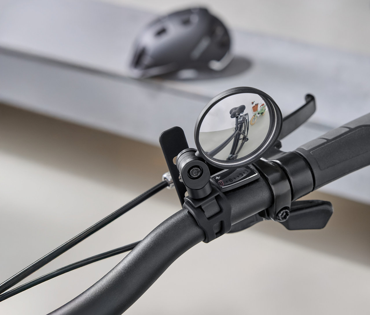 Nahaufnahme eines Fahrradlenkers mit einem runden Rückspiegel, der den dahinterliegenden Weg reflektiert. Im Hintergrund ist ein grauer Fahrradhelm zu sehen.