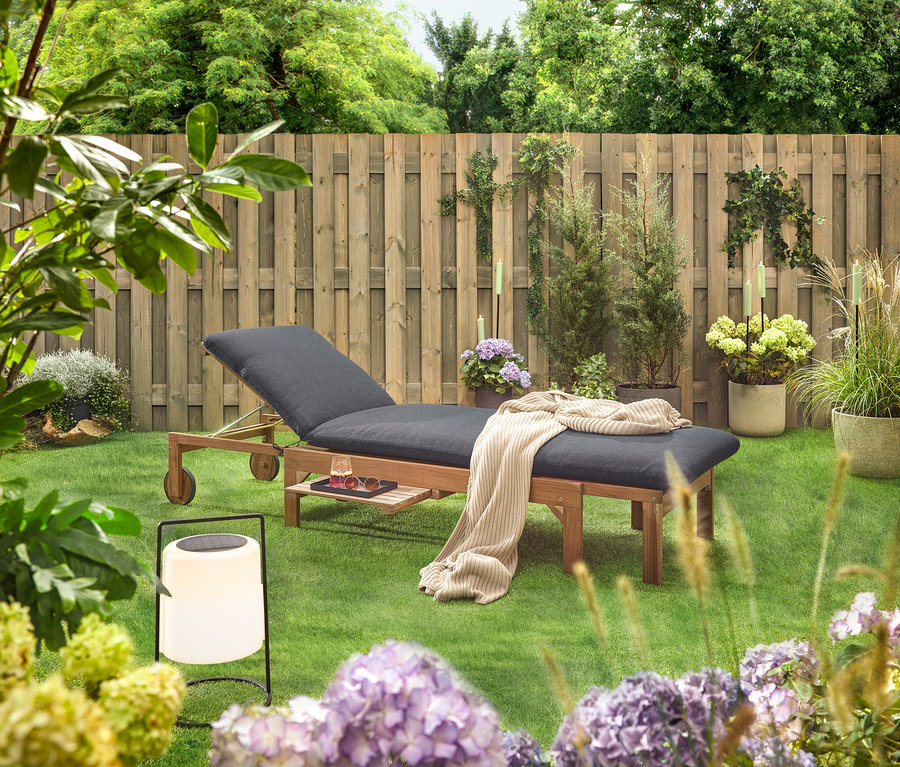 Gartenszene mit einer hölzernen Liege mit schwarzer Polsterung, einer Decke, einem Getränk, einer Sonnenbrille und einer Laterne auf einem Rasen vor einem Holzzaun.