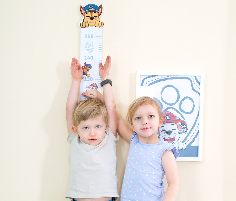 Zwei Kinder, ein Junge und ein Mädchen, stehen vor einer Wand mit einem Paw Patrol Größenmessgerät und einem gerahmten Bild.