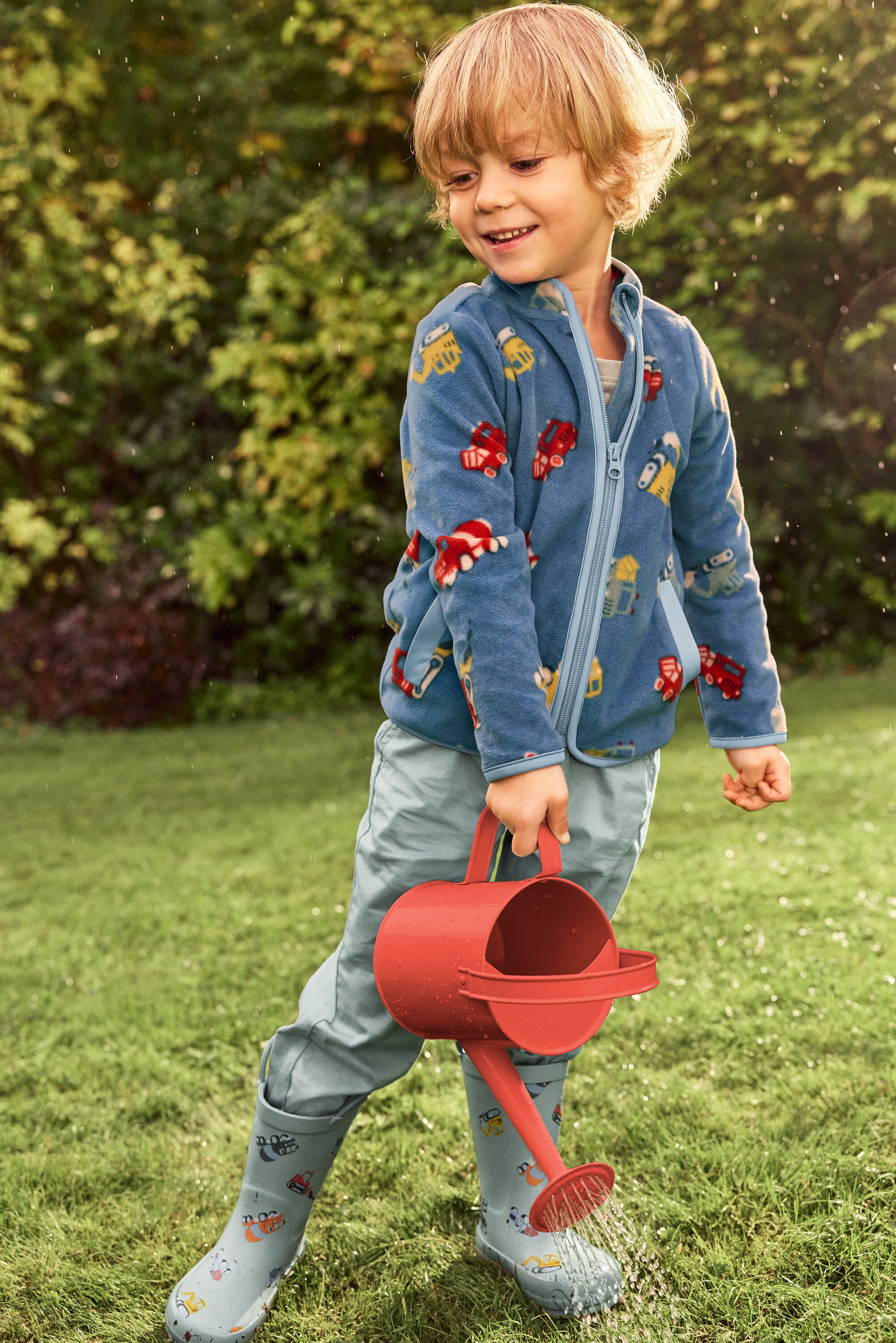 Ein lächelnder Junge mit blonden Haaren gießt mit einer roten Gießkanne Wasser auf eine grüne Wiese. Er trägt eine blaue Jacke mit Fahrzeugmotiven und graue Gummistiefel mit Fahrzeugen.