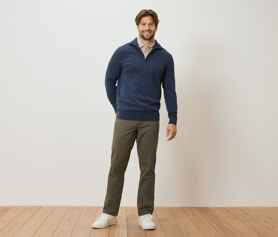 Ein Mann steht mit den Händen in den Hosentaschen vor einer Wand. Er trägt einen marineblauen Pullover, eine hellbraune Bluse, khakifarbene Hosen und weiße Turnschuhe.