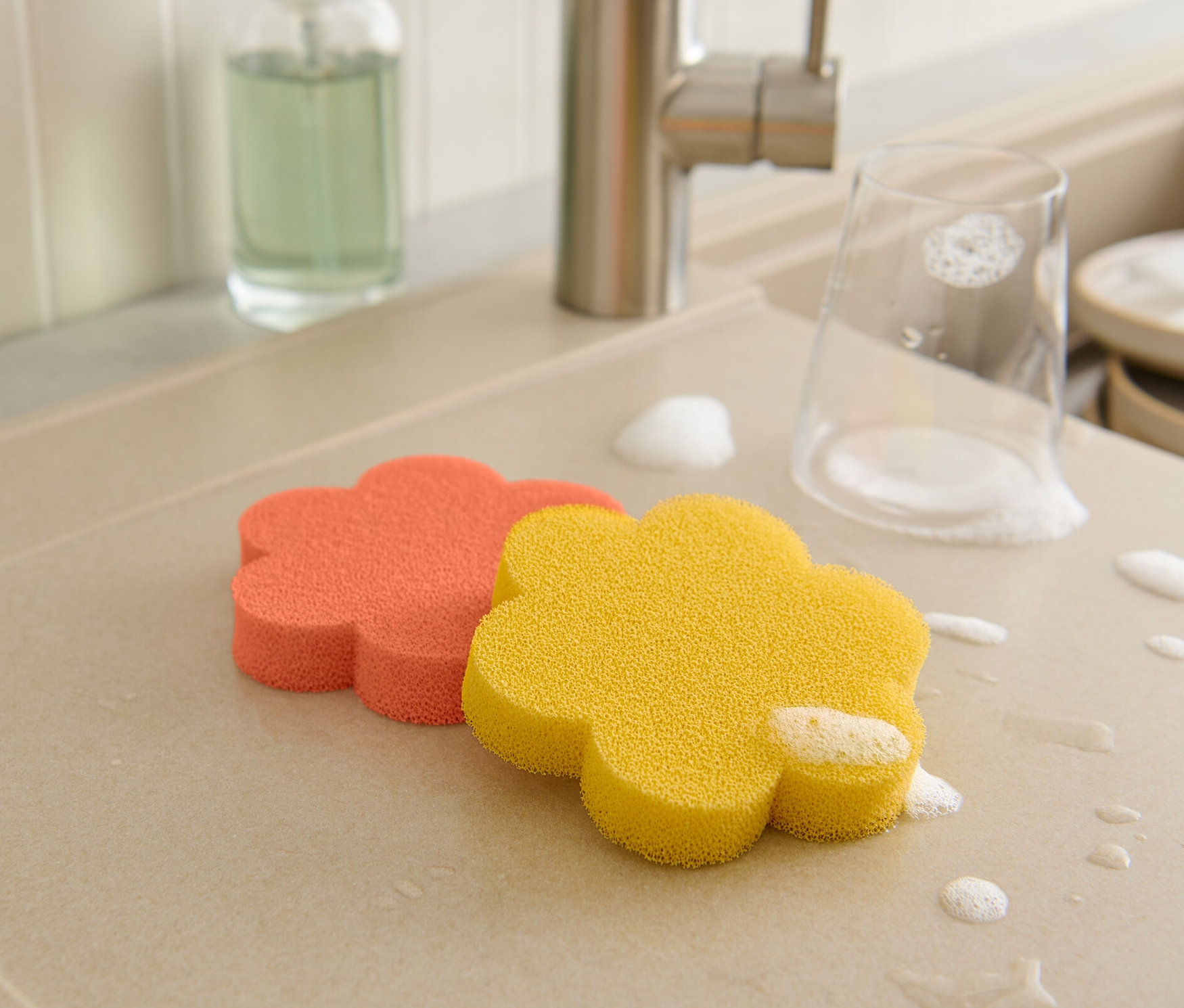 Auf der Abtropffläche liegen zwei blumenförmige Silikonschwämme, ein Glas und Geschirr sind mit Schaum bedeckt.