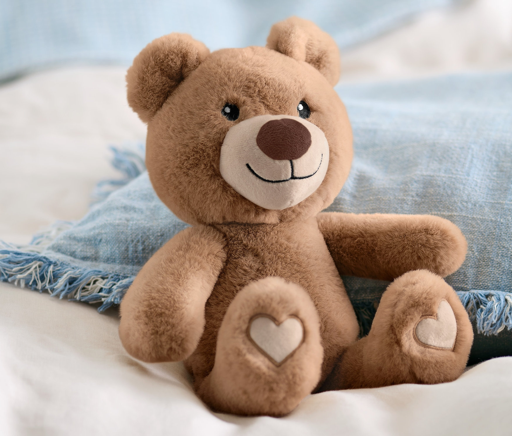 Ein braunes Plüschtier »Bär« sitzt auf weißer Bettwäsche und einem blauen Kissen.