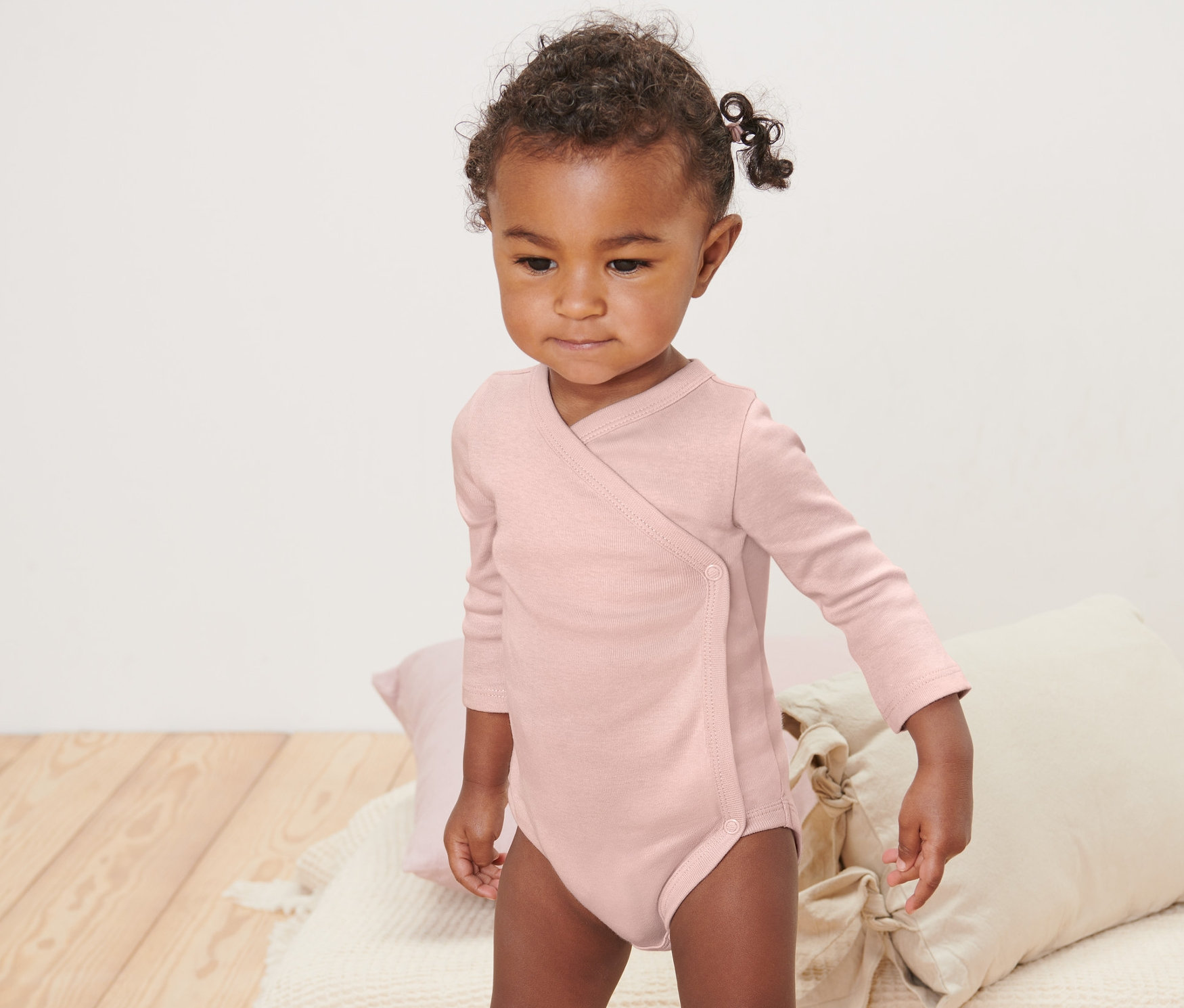 Kleinkind in einem rosa Langarmbody steht auf einer Decke. Auf der Decke liegen Kissen. 5 Baby-Bodys mit Mitwachsfunktion, rosa.