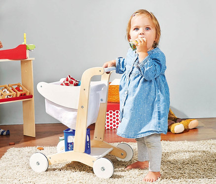 Kleinkind steht an einem weißen Pinolino Einkaufswagen »Erna« und isst einen Keks.