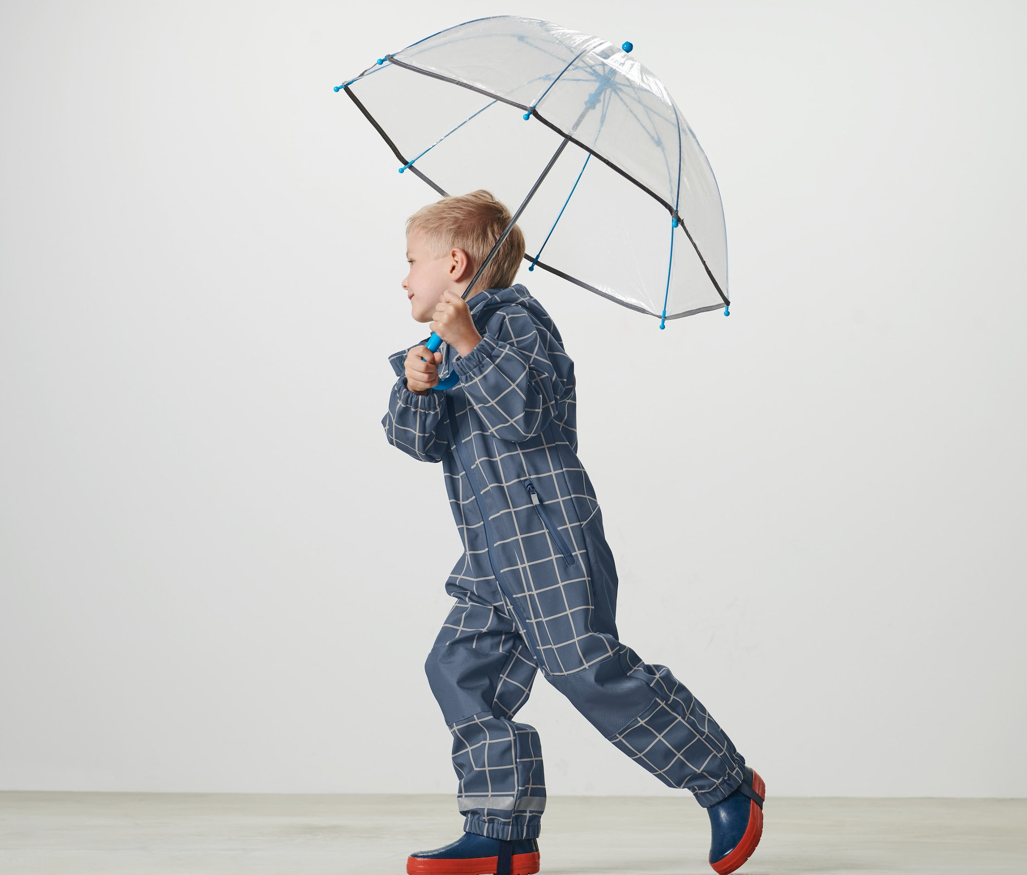 Junge trägt einen karierten Kleinkind-Softshell-Einteiler und hält einen durchsichtigen Regenschirm.