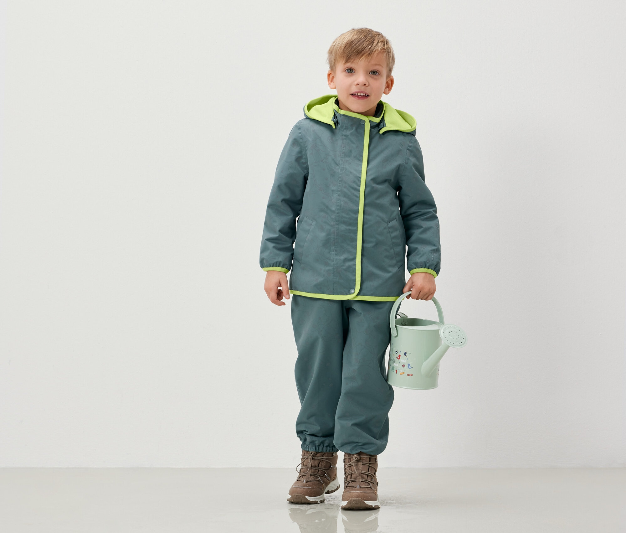 Ein Junge in Jacke und Hose hält eine hellgrüne Gießkanne.
